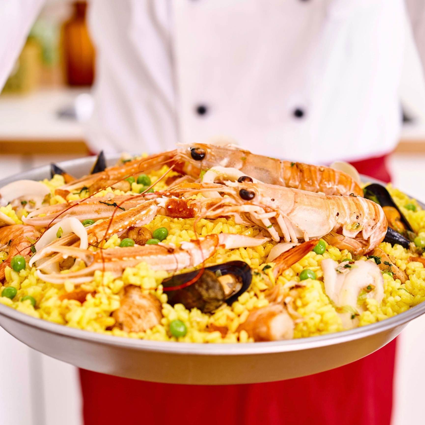 A chef presents a pan of paella with shrimp, mussels, and peas.