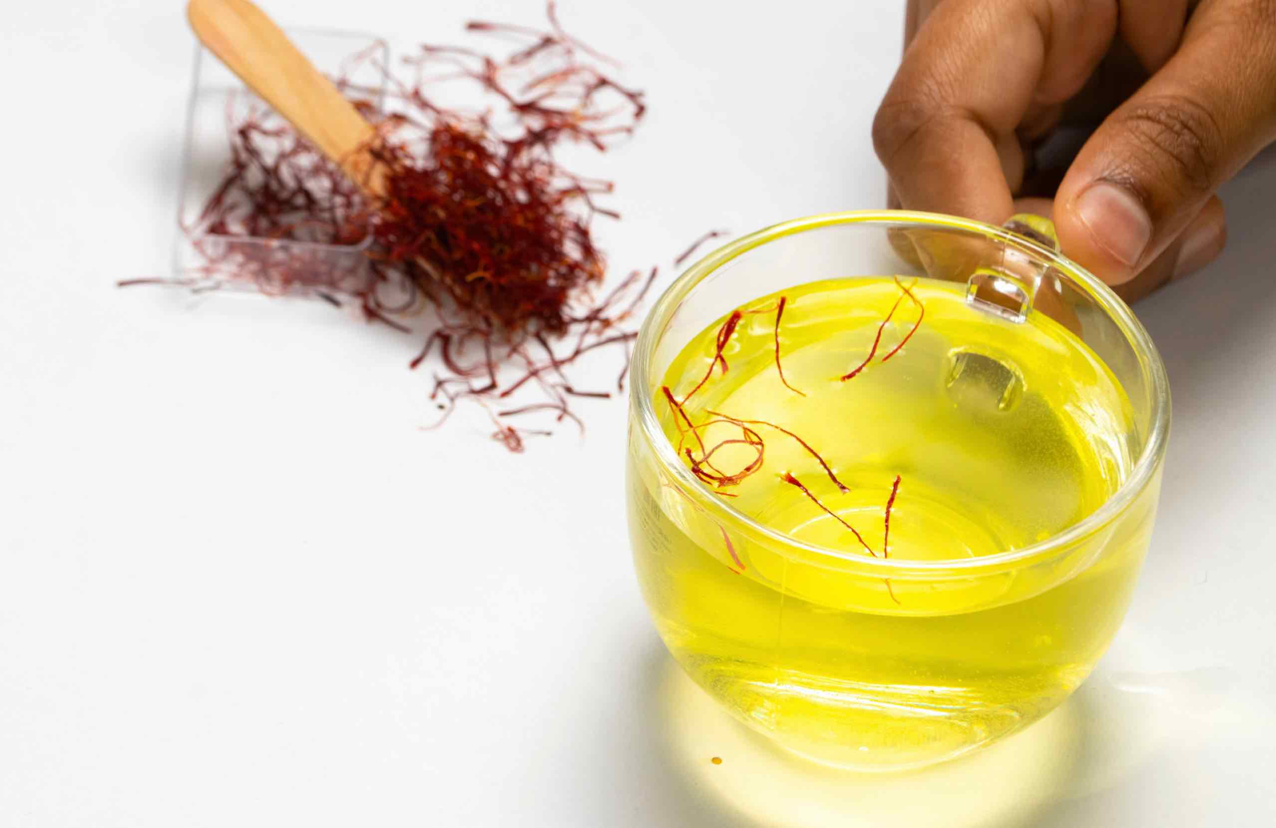 A hand holding a glass of saffron-infused liquid next to a pile of saffron threads and a wooden spoon.